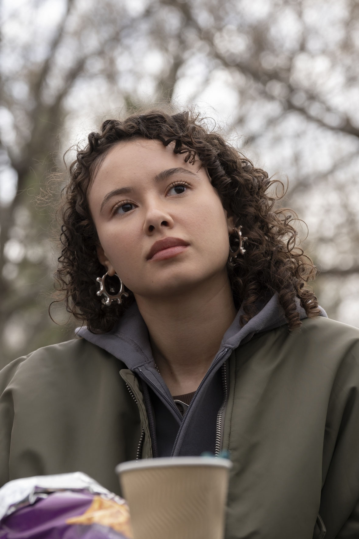 In an image from Goosebumps: The Vanishing, a photo of Alex Diaz (Francesca Noel) from the shoulders up is seen. She is standing in front of a leafless tree and looking to the right of the frame. Her head is slightly tilted and there is an out-of-focus coffee cup and chip bag in front of her.