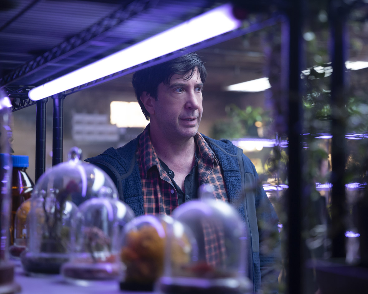 In an image from Goosebumps: The Vanishing, Anthony Brewer (David Schwimmer) stands behind shelves of bell jars containing out of focus items. He is lit with purple lighting and looking towards the right of the frame.