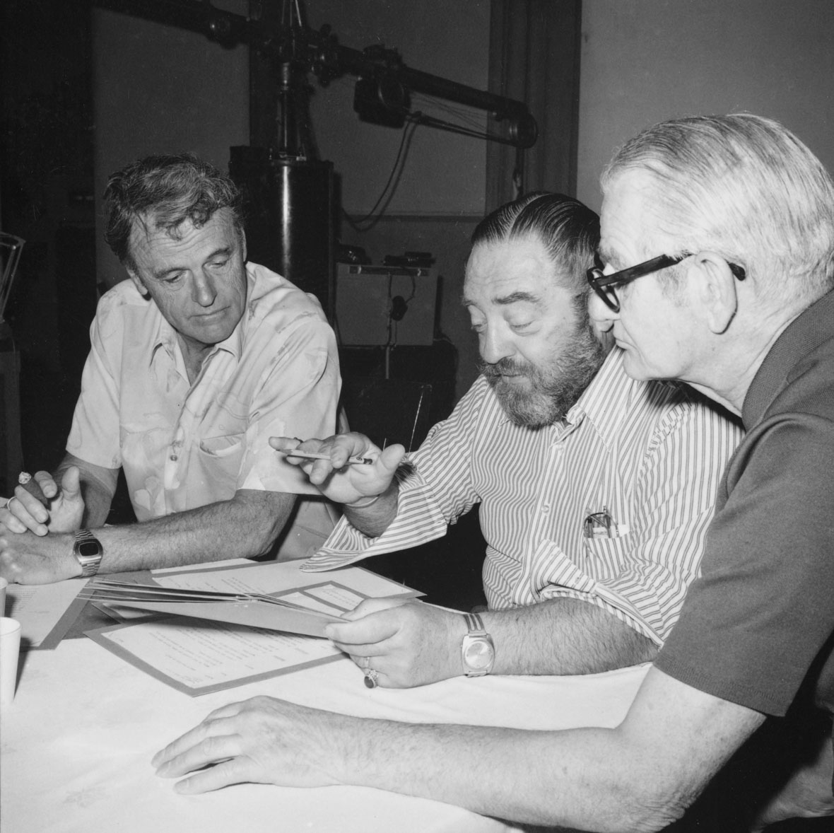 Disney Legend Woolie Reitherman, left, and Disney story artist Larry Clemmons, right, sit on either side of actor Sebastian Cabot at a table covered with papers. Cabot, who was the voice of the narrator in Disney’s Winnie the Pooh films, is holding what appears to be a script in one hand and is gesturing with the other.