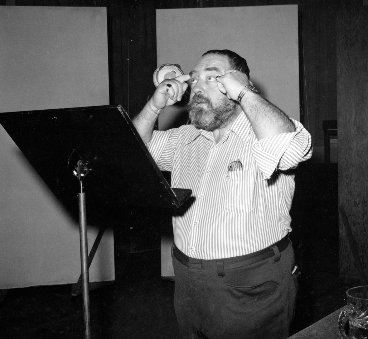 A black-and-white image of actor Sebastian Cabot in a recording booth, standing behind a script stand. His arms are raised with his index fingers touching each of his temples and his eyes are cast toward the ceiling.