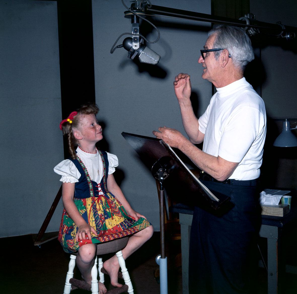 A color image of child actor Dori Whitaker in a recording booth, seated on a four-legged stool. She is looking up and smiling at Larry Clemmons, who is standing in front of her, behind a script stand. His arms are raised as if he is giving direction. Dori wears a colorful German folk skirt and white blouse and has a red ribbon in her hair, which has been shaped into pigtails.