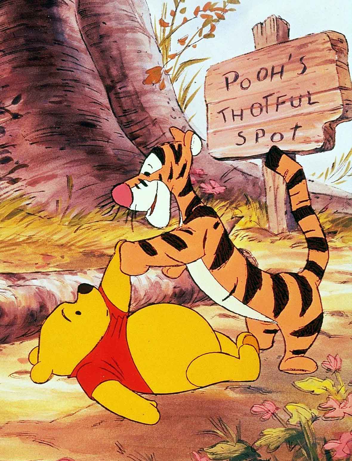 In an image from Winnie the Pooh and Tigger Too, Pooh lies on the ground, his left arm extended as he shakes hands with Tigger, who is standing at Pooh’s feet. Pooh lies next to a log and behind them is the base of a tree and a sign reading, Pooh’s Thoughtful Spot, with the word “thoughtful” misspelled.