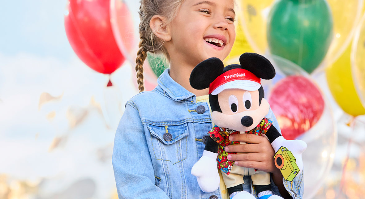 A young girl at Disneyland Park smiles with multicolored balloons behind her. She is holding a Mickey Mouse plush toy. Mickey is dressed for a day at Disneyland with a Disneyland visor, colorful short-sleeved collared button-down shirt and beige shorts.