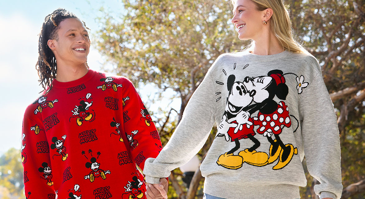 A man and woman look at each other and hold hands in an outdoor setting. The man is wearing a red Mickey Mouse sweatshirt with several images of Mickey Mouse in different action scenes adorning the sweatshirt. The Mickey Mouse logo is also featured in different areas of the sweatshirt. The woman is wearing a gray sweatshirt feature Minnie Mouse giving Mickey a kiss