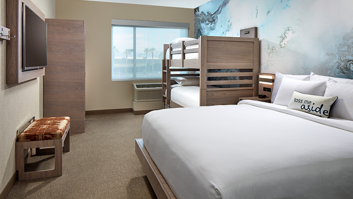 An example of the hotel&rsquo;s rooms, with a clean and cozy design&mdash;featuring a king-sized bed adorned with white linens and a decorative pillow that reads &ldquo;Toss me aside.&rdquo; Behind the bed is an abstract blue wall mural. The room also includes a wooden bunk bed, a wall-mounted TV, a small bench, and a window with natural light illuminating the space.