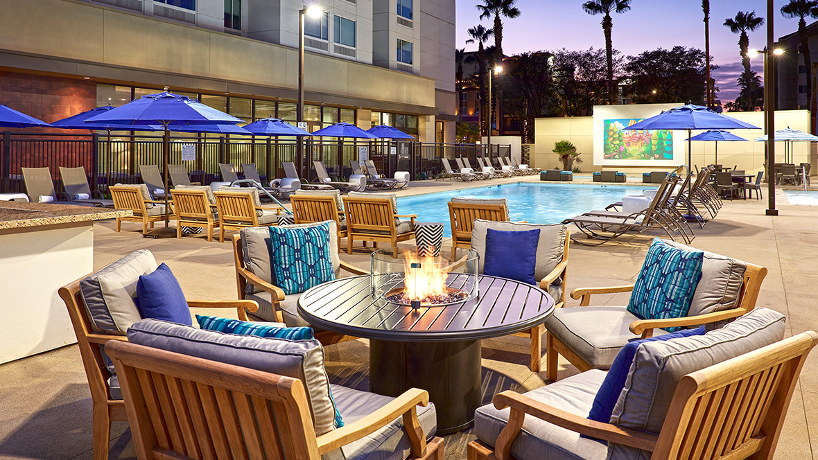 Outdoor pool area at dusk&mdash;featuring seating arrangement with wooden chairs and plush cushions around a lit fire pit. Behind the fire pit, blue umbrellas and wicker lounge chairs line the pool, with a colorful mural in the background. Palm trees dot a twilight sky to the right.
