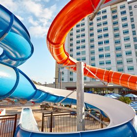 A photo of the pool area at Cambria Hotel & Suites Anaheim Resort. The vibrant outdoor scene features two colorful water slides, one blue and one orange, curving toward lounge chairs and striped umbrellas. A modern-style hotel building can be seen past the slides; at its top is a sign reading “Cambria.” To the building’s left, a sunny, blue sky.