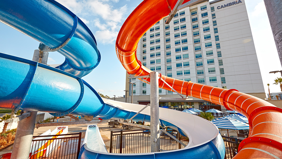 A photo of the pool area at Cambria Hotel & Suites Anaheim Resort. The vibrant outdoor scene features two colorful water slides, one blue and one orange, curving toward lounge chairs and striped umbrellas. A modern-style hotel building can be seen past the slides; at its top is a sign reading “Cambria.” To the building’s left, a sunny, blue sky.