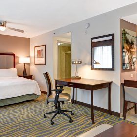 An example room at the Homewood Suites by Hilton Lake Buena Vista. A large bed sits between two fully lit nightstands. Opposite the bed, on the right, is a doorway into the bathroom as well as a rectangular desk with an office chair. To the right of the desk is a dining table with several chairs and kitchenet; in the kitchenet is a sink, a microwave, and dark brown cabinets.