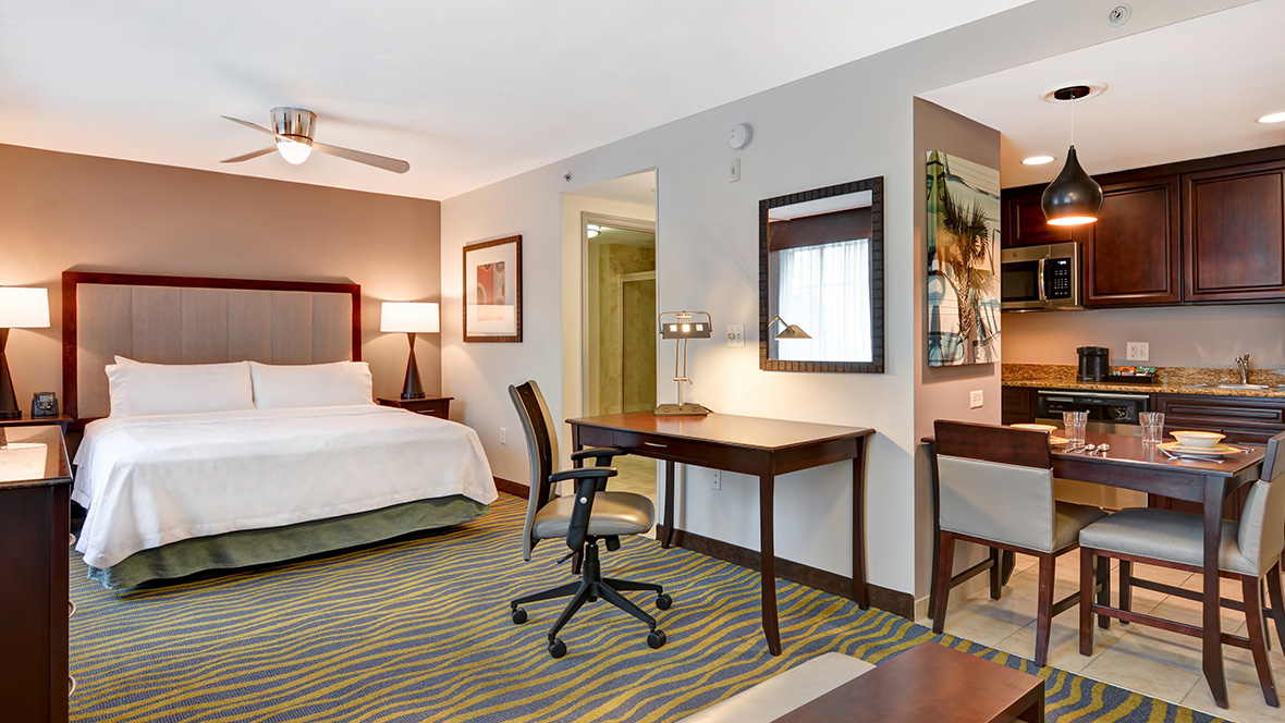 An example room at the Homewood Suites by Hilton Lake Buena Vista. A large bed sits between two fully lit nightstands. Opposite the bed, on the right, is a doorway into the bathroom as well as a rectangular desk with an office chair. To the right of the desk is a dining table with several chairs and kitchenet; in the kitchenet is a sink, a microwave, and dark brown cabinets.