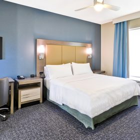 A photo of a sample room at Homewood Suites by Hilton Orlando Theme Parks. A queen-sized bed is at the middle-right of the room, with a white duvet and pillows. On either side of the yellow headboard are two lit lamps. To the left of the bed is a large window with a wall air conditioning unit at its base. Two light-blue drapes frame the window. To the left of the bed is a nightstand; to the left of the nightstand is a desk with an office chair.