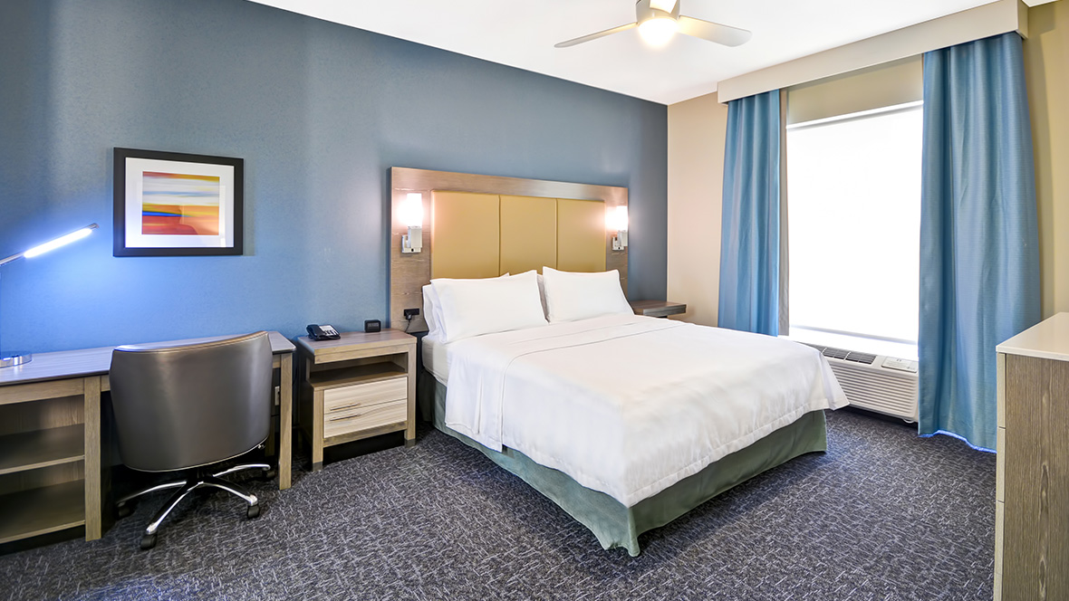 A photo of a sample room at Homewood Suites by Hilton Orlando Theme Parks. A queen-sized bed is at the middle-right of the room, with a white duvet and pillows. On either side of the yellow headboard are two lit lamps. To the left of the bed is a large window with a wall air conditioning unit at its base. Two light-blue drapes frame the window. To the left of the bed is a nightstand; to the left of the nightstand is a desk with an office chair.