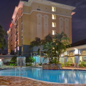 The Hilton Garden Inn Lake Buena Vista/Orlando sits brightly lit against the night sky. In front of the yellow hotel is the pool area. The pool is a turquoise blue color with the hotel lights reflected within the water. Multiple railings and steps are connected to the pool for access. Surrounding the pool are assorted lounge chairs and palm trees. Next to the pool is a small blue building with the lights on inside.