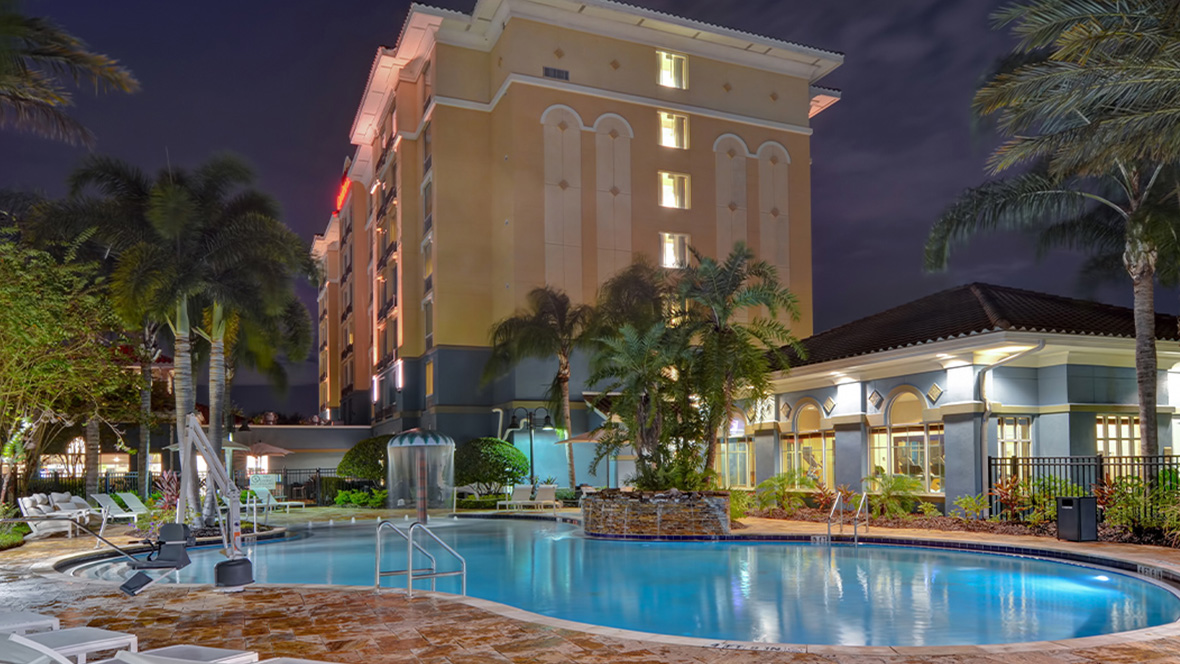 The Hilton Garden Inn Lake Buena Vista/Orlando sits brightly lit against the night sky. In front of the yellow hotel is the pool area. The pool is a turquoise blue color with the hotel lights reflected within the water. Multiple railings and steps are connected to the pool for access. Surrounding the pool are assorted lounge chairs and palm trees. Next to the pool is a small blue building with the lights on inside.