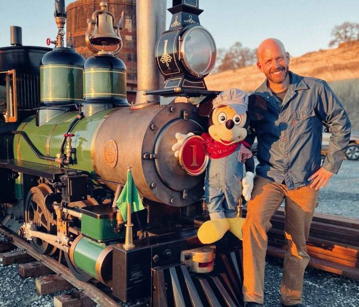 A photograph on a train track, on the left side is the front of a train. Bret Iwan (artist and voice of Mickey Mouse) is standing to the right of train, holding a plush Mickey Mouse dressed as a train engineer. 