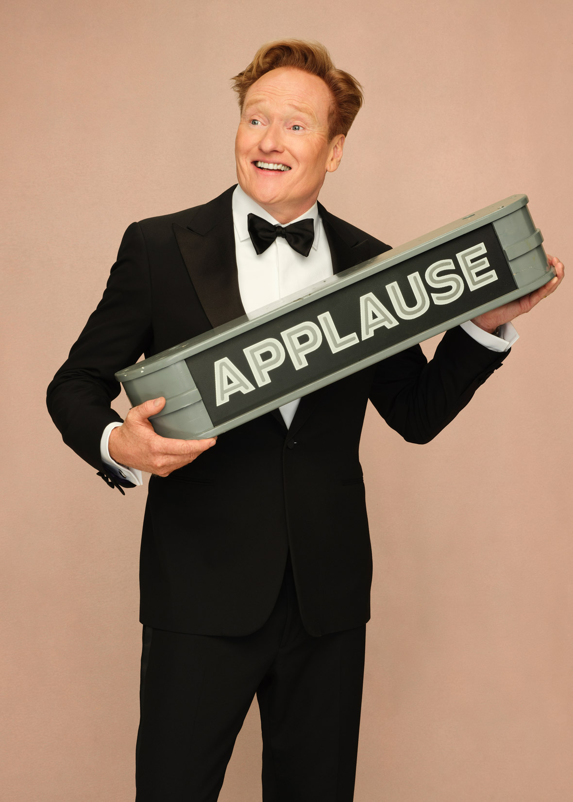 Conan O'Brien wears a tuxedo and holds an applause sign.