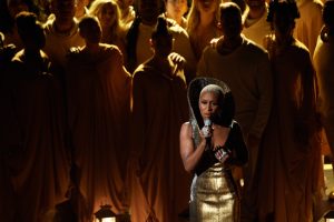 Cynthia Erivo performs during the 92nd Oscars in 2020. She is wearing a form-fitting gold gown with an oversized collar and stands in front of a choir wearing gold robes.