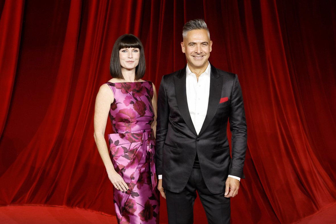 Katy Mullan and Raj Kapoor wear eveningwear and stand in front of a red curtain.