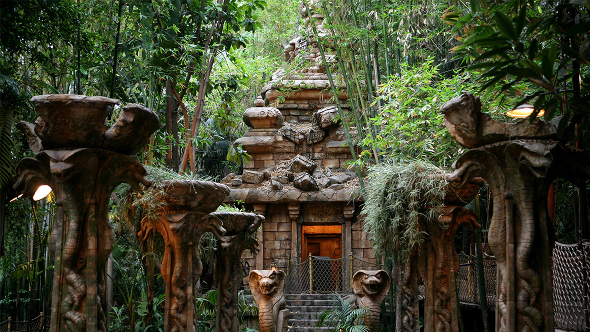 Outside queue of the Indiana Jones Adventure attraction at the Disneyland Resort. Green foliage and tall columns are seen in front of a brown stone temple, guarded by two snake statues.