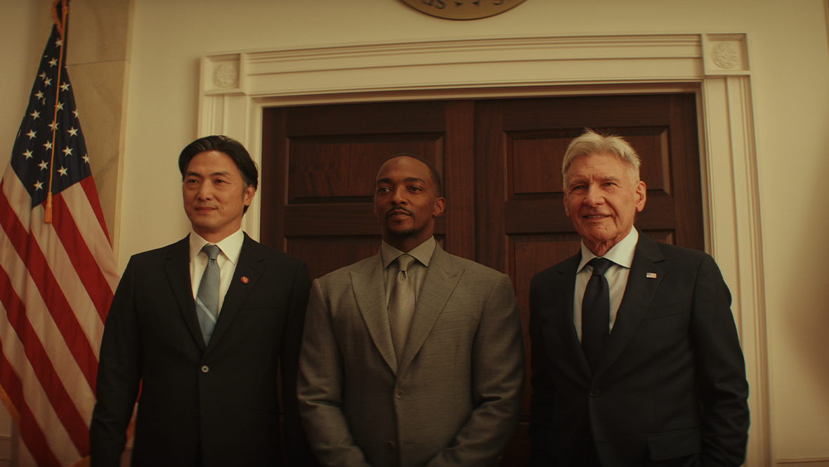 Harrison Ford is U.S. President Thaddeus “Thunderbolt” Ross in this image from Captain America: Brave New World. Ross, at the center of the photo, is standing with Sam Wilson, aka Captain America (played by Anthony Mackie), to the right, and, on the left, Prime Minister Ozaki (played by Takehiro Hira). All three are in suits and ties, facing forward with serious expressions on their faces. Behind them is a closed doorway and a wall, presumably within the White House, and they are flanked by two full-size flags – the U.S. flag on the left and the flag representing the U.S. President on the right. A dispay of pink cherry blossoms is on the far left of the photo.