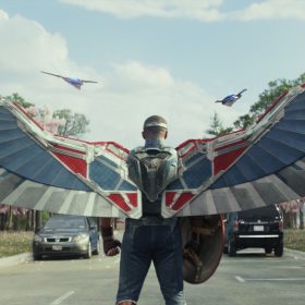 Sam Wilson in Captain America: Brave New World. He is turned away and wearing outstretched grey, red, and blue mechanical wings while holding the Captain America shield. There are two cars and cherry blossoms trees in the background of the image.