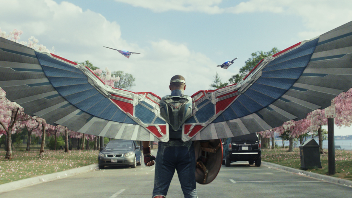 Sam Wilson in Captain America: Brave New World. He is turned away and wearing outstretched grey, red, and blue mechanical wings while holding the Captain America shield. There are two cars and cherry blossoms trees in the background of the image.