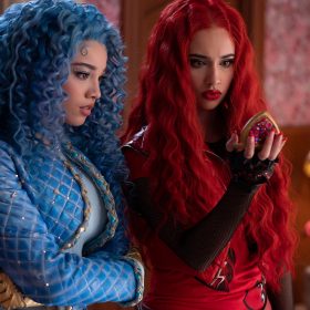 Chloe, played by Malia Baker, stands next to Princess Red, played by Kylie Cantrall, in a scene from Disney Branded Television's Descendants: The Rise of Red. Chloe has curly blue hair and is wearing a blue, quilted, bedazzled jacket, a blue shirt, and blue pants. Red has wavy red hair and is wearing a red leather vest over a red T-shirt and black, long-sleeved fishnet top. Red is holding a jewel-encrusted, heart-shaped pocket watch.