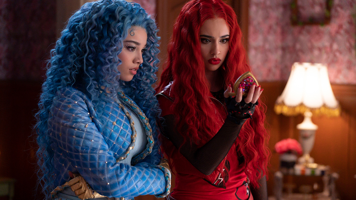 Chloe, played by Malia Baker, stands next to Princess Red, played by Kylie Cantrall, in a scene from Disney Branded Television's Descendants: The Rise of Red. Chloe has curly blue hair and is wearing a blue, quilted, bedazzled jacket, a blue shirt, and blue pants. Red has wavy red hair and is wearing a red leather vest over a red T-shirt and black, long-sleeved fishnet top. Red is holding a jewel-encrusted, heart-shaped pocket watch.