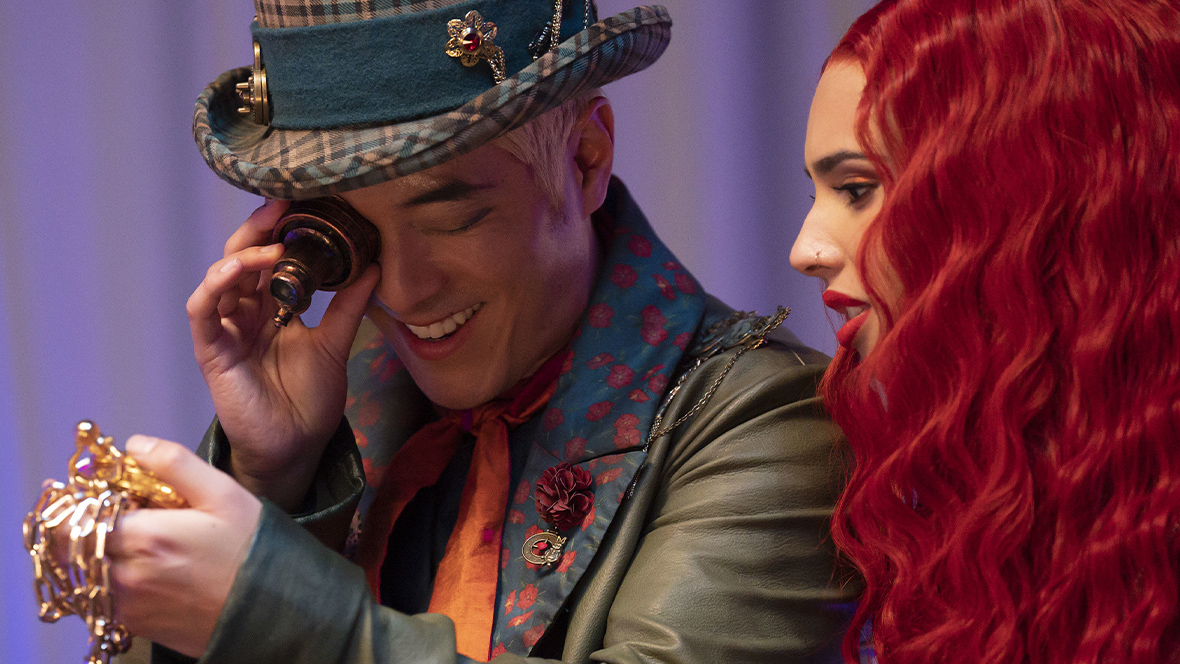 In an image from Descendants: The Rise of Red, Maddox Hatter, played by Leonardo Nam, is wearing a plaid top hat, a green leather jacket, and an orange scarf. He uses a magnifying glass to inspect a gold pocket watch. Princess Red, who has wavy red hair, is looking over Maddox Hatter's left shoulder.