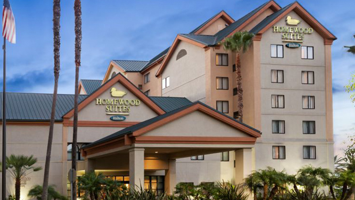 The Homewood Suites by Hilton Anaheim Maingate stands in front of the afternoon blue sky. The hotel is made up of several towers featuring triangle rooftops; the Homewood Suites logo is seen on the top of both the small and large tower. Small palm trees are seen near the front of the hotel.