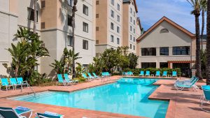 Aqua blue chairs line the pool area of the Homewood Suites by Hilton Anaheim Maingate. Behind the chairs are green palm trees of several different sizes; a table off to the right is set up for guests to enjoy a meal in the Southern California sunshine. To the left of the pool are the towering buildings that make up the rooms of the hotel. The pool is a blue turquoise color.