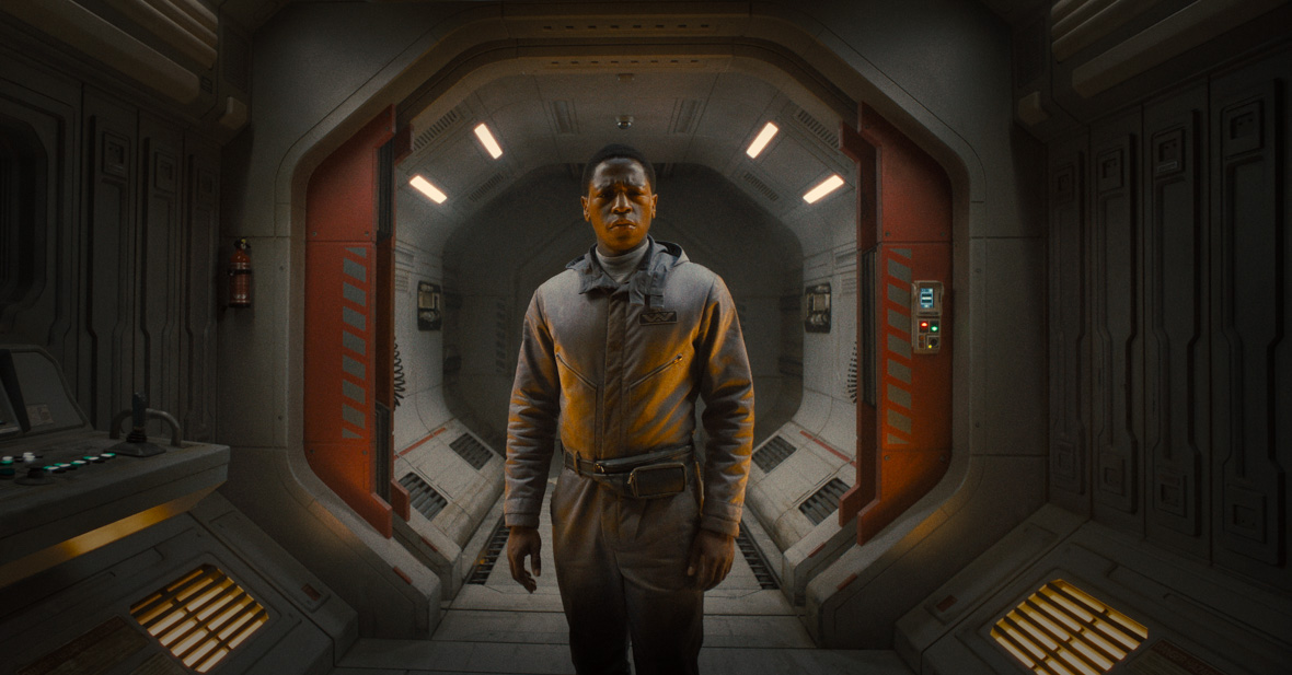 David Jonsson as Andy in Alien: Romulus; he’s wearing a gray jumpsuit and looking concerned. He is standing in a gray, futuristic-looking corridor on a spaceship and is in front of a pair of open orange door