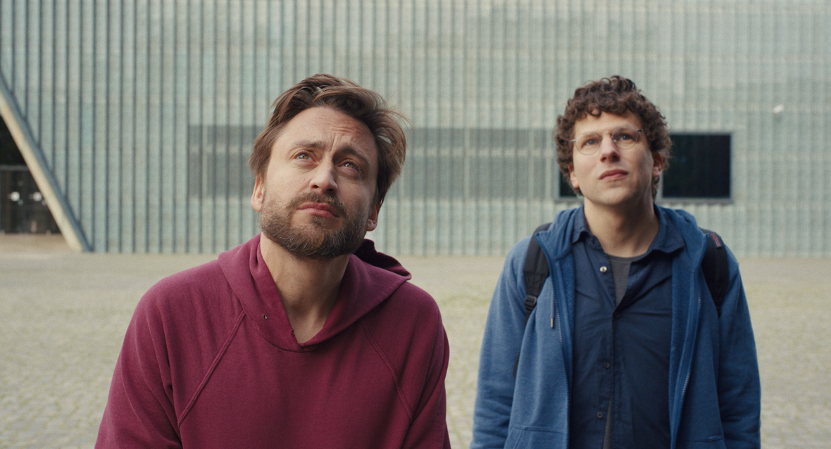 Jesse Eisenberg as David Kaplan and Kieran Culkin as Benji Kaplan in A Real Pain. Eisenberg is wearing circular glasses, a blue shirt, a blue hoodie, and a black backpack. Culkin is wearing a red pullover. They are both standing in front of a grey wall and looking up into the distance.