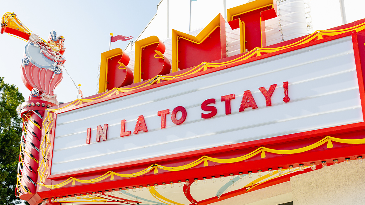 A vibrant marquee sign of the Bob Baker Marionette Theater in Los Angeles, painted in red, yellow, and white, with decorative trim and a playful, vintage aesthetic. The sign displays bold red letters spelling 'IN LA TO STAY!' The top of the marquee features a whimsical drum-playing character, enhancing the theater's nostalgic and lively charm.