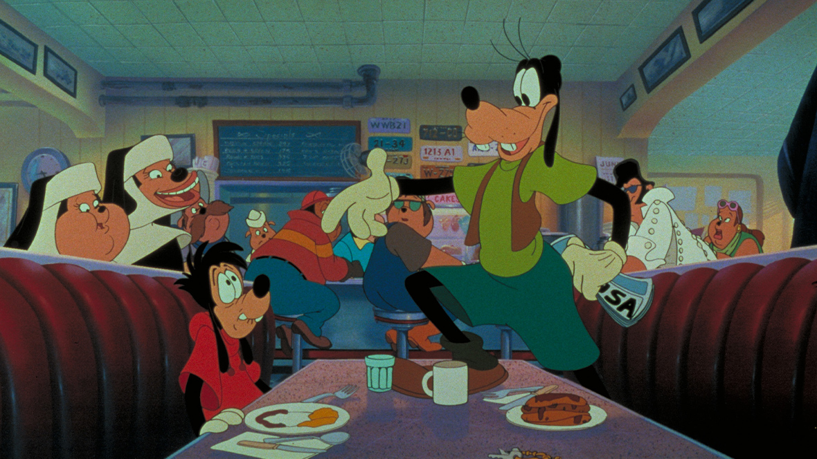 A screengrab from the film A Goofy Movie, set in a diner. On the left, Max Goof sits in a diner booth with a plate of breakfast food. To the right, Goofy is standing up to present Max their road trip map to have him be in charge of navigation. Diners in the restaurant are in the background observing this moment.