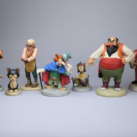 Nine colorful animator’s models from the development of the 1940 animated feature Pinocchio—left to right: Jiminy Cricket (posed pointing to the right), Pinocchio (posed pointing up), the cat Figaro, Geppetto (in a pensive pose), the fox J. Worthington Foulfellow, sidekick cat Gideon, Stromboli (pointing down with his right hand), the Coachman, and Lampwick—are displayed against a gray backdrop.