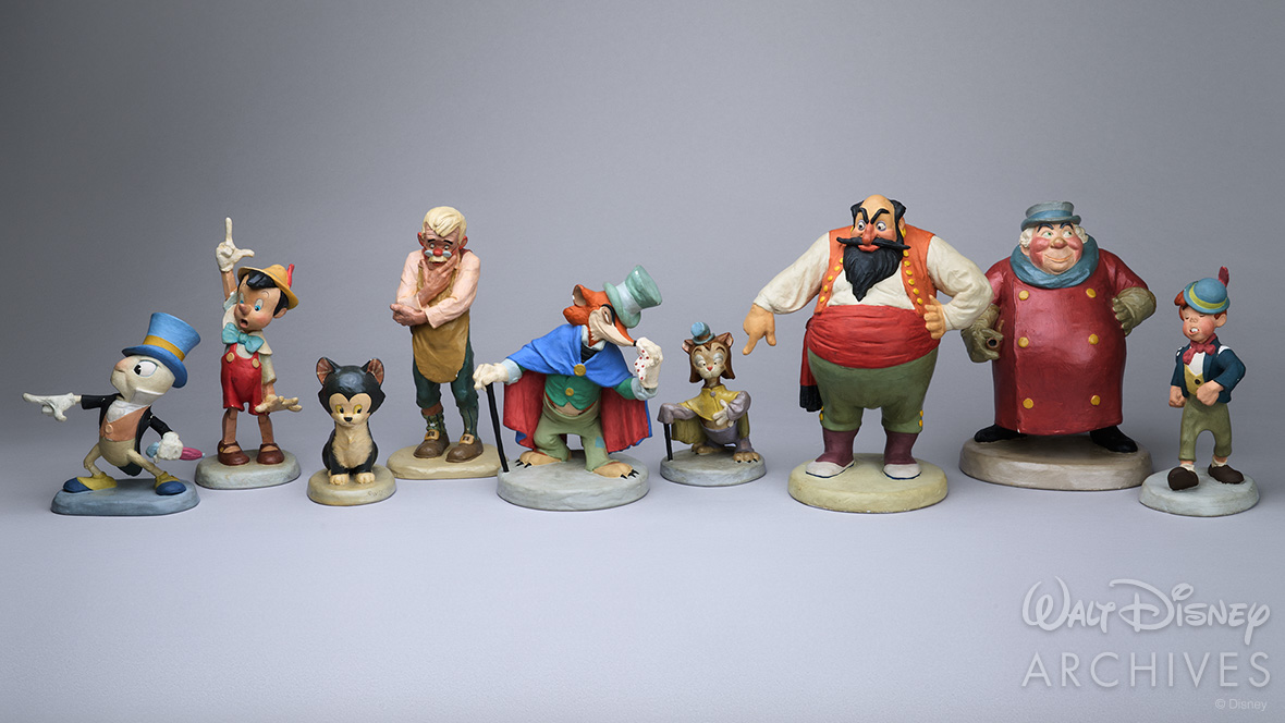 Nine colorful animator’s models from the development of the 1940 animated feature Pinocchio—left to right: Jiminy Cricket (posed pointing to the right), Pinocchio (posed pointing up), the cat Figaro, Geppetto (in a pensive pose), the fox J. Worthington Foulfellow, sidekick cat Gideon, Stromboli (pointing down with his right hand), the Coachman, and Lampwick—are displayed against a gray backdrop.