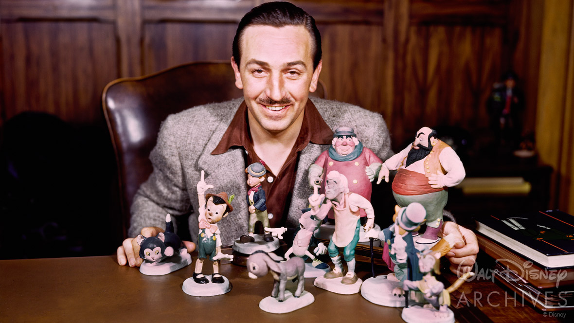 Walt Disney, dressed in a brown collared shirt and grey coat, sits at a desk behind ten animator models from the 1940 animated feature Pinocchio. 