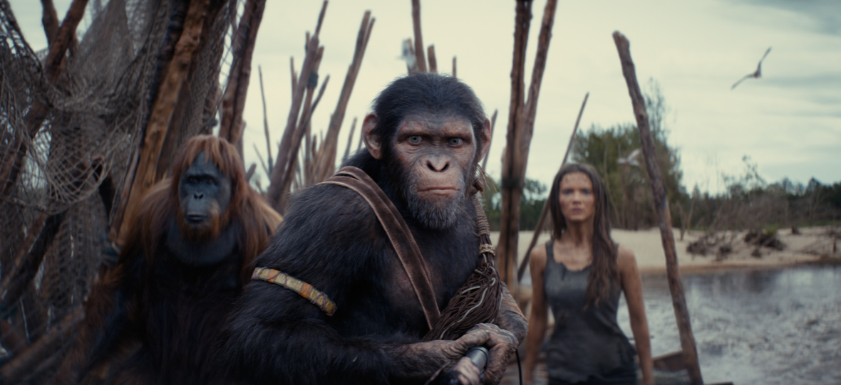 Noa, a chimpanzee from Kingdom of the Planet of the Apes, stands center while holding a spear outward. Behind him are a Raka, an orangutan, and Nova, a woman, wearing a grey shirt. They are on a raft near the bank of a river.