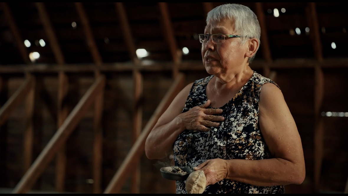 In an image from National Geographic’s Sugarcane, Charlene Belleau, an older woman with short, gray hair and glasses, wears a white and brown sleeveless blouse. She’s holding out a small dish full of herb in her left hand and her right hand is on her heart. She is standing in front of a brown background with visible wooden supports.