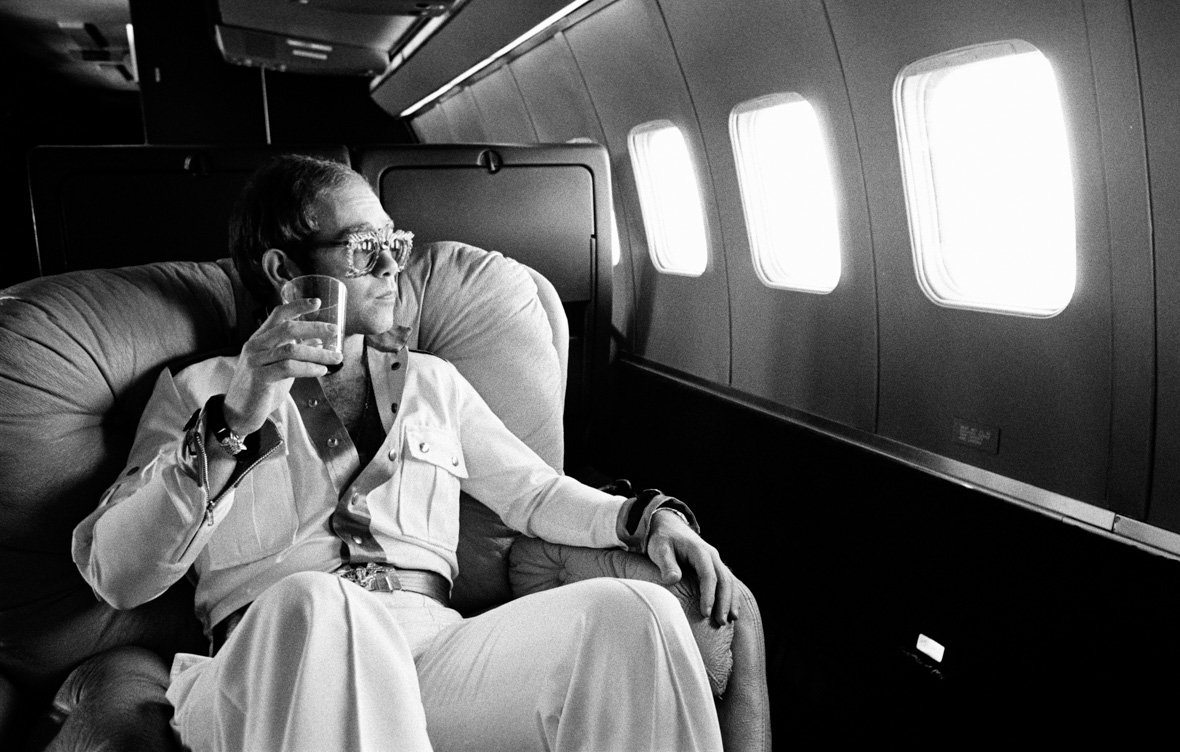 A black and white photo of Disney Legend Sir Elton John in 1974. He’s sitting on a seat in an airplane and staring out the window while holding a drinking glass, while wearing a white jumpsuit and large glasses.