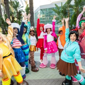 An image of a group of cosplayers, standing in front of the Anaheim Convention Center during D23: The Ultimate Disney Fan Event. They’re dressed as Vanellope von Schweetz and her fellow Sugar Rush racers from Wreck-It Ralph; their outfits are colorful and several cosplayers wear oversized hats and shoes.