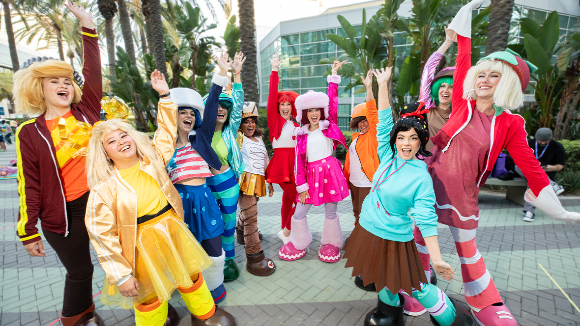 An image of a group of cosplayers, standing in front of the Anaheim Convention Center during D23: The Ultimate Disney Fan Event. They’re dressed as Vanellope von Schweetz and her fellow Sugar Rush racers from Wreck-It Ralph; their outfits are colorful and several cosplayers wear oversized hats and shoes.