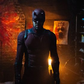 A figure dressed in Daredevil's red armored suit stands in a dimly lit room filled with graffiti-covered walls and shelves stocked with spray paint cans and tools.