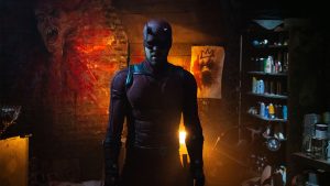 A figure dressed in Daredevil's red armored suit stands in a dimly lit room filled with graffiti-covered walls and shelves stocked with spray paint cans and tools.