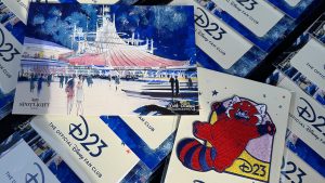 Close-up photo of the event gifts from the D23 Spotlight Series & Walt Disney Imagineering Present Space Mountain and Beyond event— a postcard of Space Mountain concept art, and a D23 Turning Red patch. The items are set on top of rows of the same gift pairings.