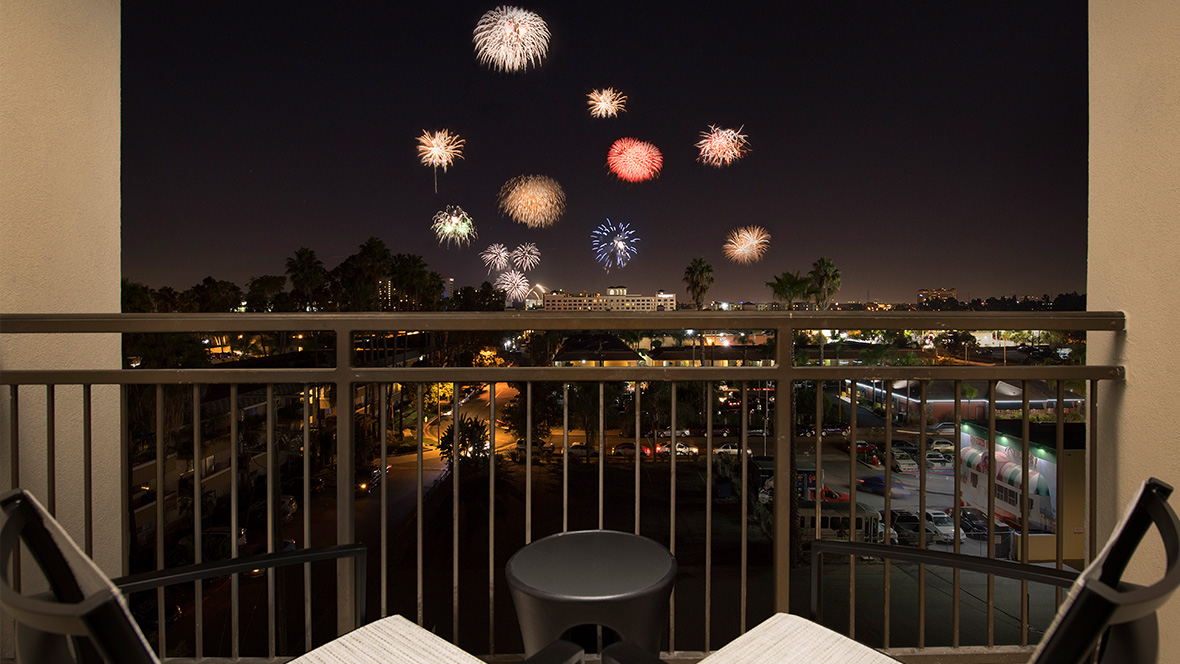 Two chairs sit out on the hotel balcony overlooking Anaheim during the nighttime. The scattered city buildings are illuminated from within; above the cityscape is a multi-colored and multi-shaped firework show that lights up the sky.