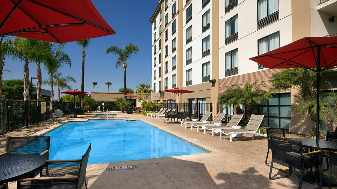 A crisp below-ground blue pool rests next to the Hampton Inn & Suites Anaheim, Garden Grove, CA. Within the patio of the pool are two round tables each with four chairs. Umbrellas are installed at the center of each table. Along the edge of the pool are several lounge chairs, extended out for relaxing.