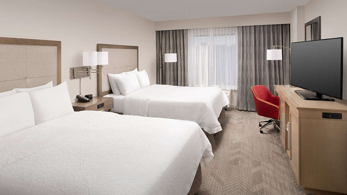 A representative room at the Hampton Inn & Suites Anaheim, Garden Grove, CA. Two queen-sized beds are separated by a nightstand, with a lamp extending from the wall. Opposite the beds is a small wooden dresser with a flatscreen TV sitting on top. Beyond the dresser, on the right, is a red rolling office chair with another extended lamp above it. The floor of the room is a patterned tan carpet.