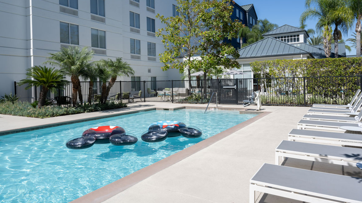 The below-ground pool of the Hilton Garden Inn Anaheim in Garden Grove sits just beyond the hotel. The sky is blue and various trees and shrubbery are scattered near the pool water. Multiple lounge benches rest next to the edge of the pool, ready for guests looking to relax. Two inner tubes in the shape of Mickey Mouse float along the clear turquoise pool water.